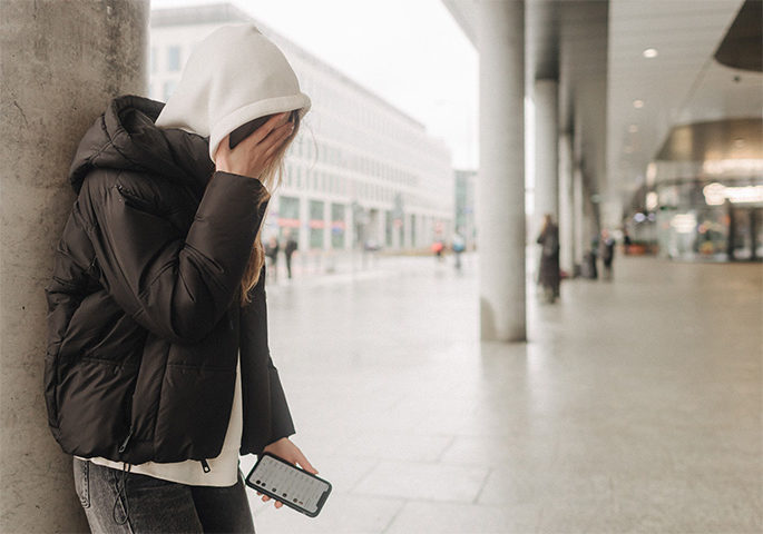 Junge Person im Hoodie mit Handy in der Hand verdeckt ihr Gesicht.