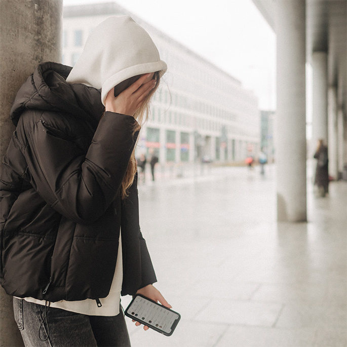 Junge Person im Hoodie mit Handy in der Hand verdeckt ihr Gesicht.