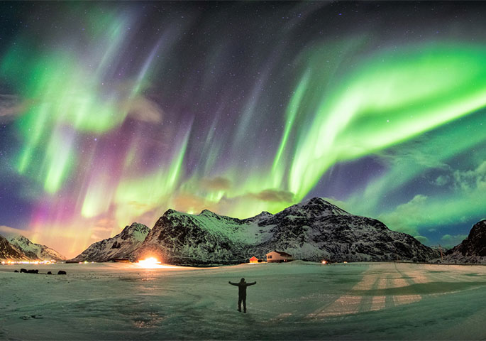 Person mit ausgestreckten Armen auf einem Eisfeld bewundert Polarlichter am Himmel vor einer Bergkulisse.