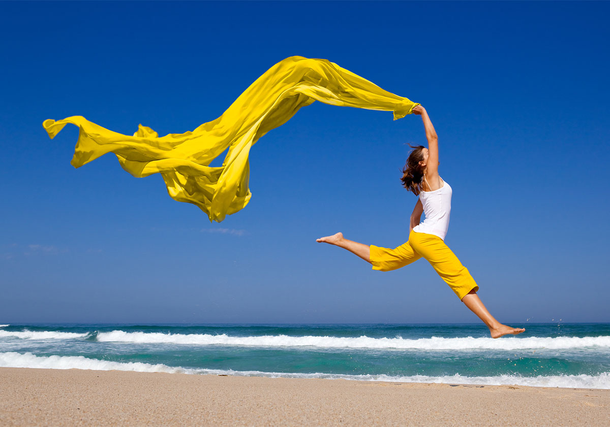 Frau mit gelbem Tuch im Wind springt am Strand.