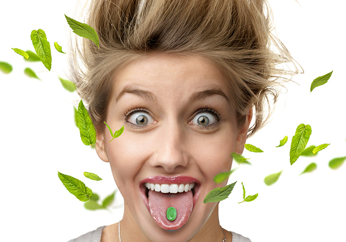 Frau mit aufgerissenen Augen und grünem Bonbon auf der Zunge von dem Minzblätter wegfliegen, stehen die Haare hoch.