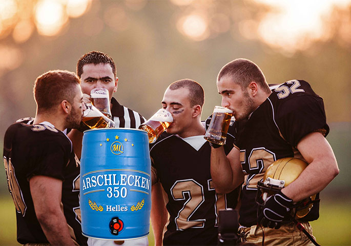 Footballspieler trinken Bier. Im Vordergrund ein kleines Bierfass mit Aufschrift Arschlecken 3 50.