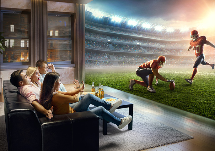 Freunde schauen auf Couch Football. Statt Fernseher sieht es so aus, als wären sie direkt im Stadion.